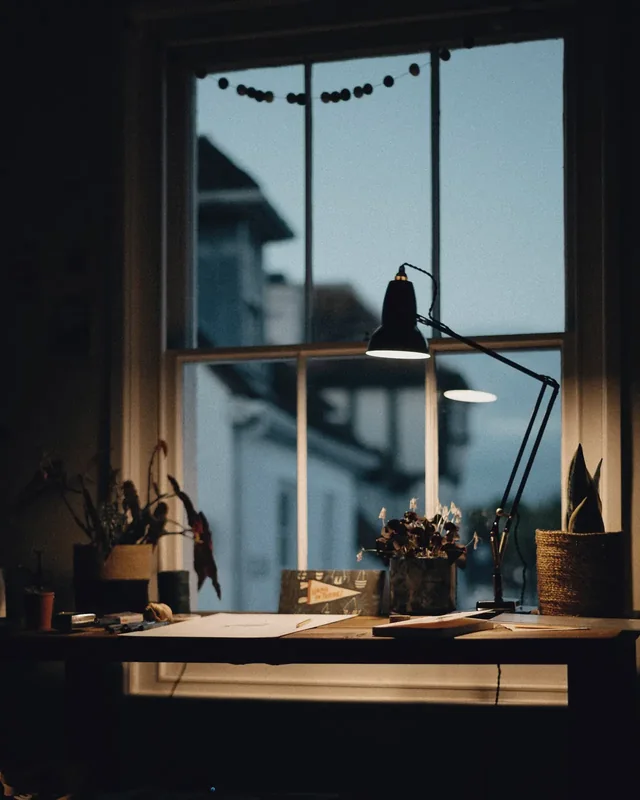 Un bureau près de la fenêtre au crépuscule, la lampe allumée.